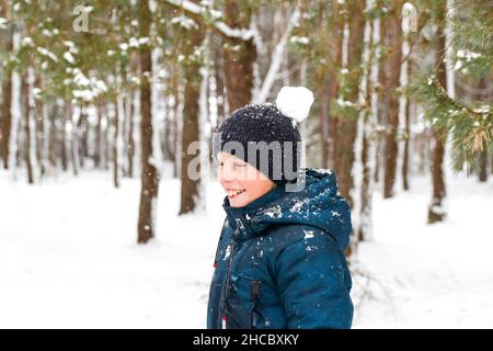 Unschärfe lächelndes Kind Junge im Winterwald im Schneesturm mit Schneeball auf dem Kopf. Lustiges Kind in der Nähe von Baum auf verschneiten Winterhintergrund stehen. Aktive Ausgaben Stockfoto