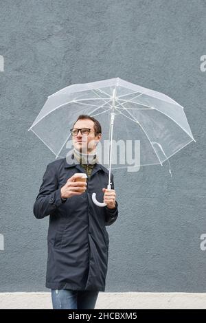 Vertikale mittellange Aufnahme eines eleganten kaukasischen Mannes mit Brillen, die unter einem Regenschirm stehen und eine Tasse Kaffee wegschauen Stockfoto