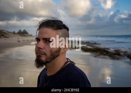 Weißer Mann zwischen 25 und 30 Jahren mit einem modernen Haarschnitt und einer misstrauischen Haltung am Meeresrand. Stockfoto