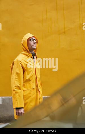 Vertikale mittellange Aufnahme eines reifen kaukasischen Mannes mit gelbem Regenmantel, der im Regen läuft und wegschaut Stockfoto