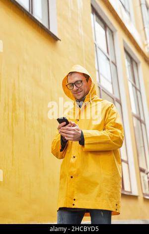 Vertikale Low-Angle-Aufnahme eines reifen kaukasischen Mannes mit gelbem Regenmantel, der mit dem Smartphone im Regen läuft Stockfoto