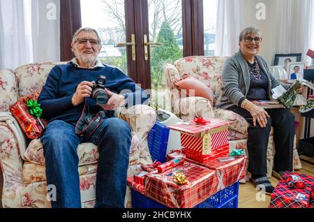 Ein Rentnerpaar sitzt am Weihnachtsmorgen in Sesseln mit Geschenken zum Öffnen, aber schaut glücklich zu, wie die Familie unsere von geschossenen offenen Geschenken zuerst schaut. Stockfoto