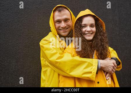 Horizontales Porträt des kaukasischen Mannes und der verliebten Frau in gelben Regenmänteln, die sich umarmen und die Kamera betrachten Stockfoto