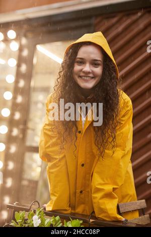 Vertikale mittlere Aufnahme einer attraktiven kaukasischen Frau mit gelbem Regenmantel, die im Regen steht und lächelt vor der Kamera Stockfoto