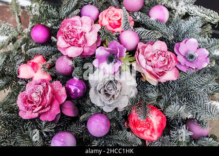 Weihnachtliche Blumenarrangements von trockenen Tannenzweigen, künstlichen Anemonen und Rosen in Himbeer- und Purpurtönen, bedeckt mit Schnee Stockfoto