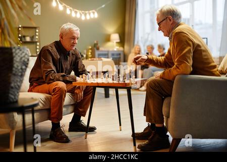 Zwei ältere kaukasische Männer, die im Wohnzimmer auf Sesseln vor einander sitzen und Schach spielen, während drei ältere Frauen auf dem Hintergrund sitzen Stockfoto