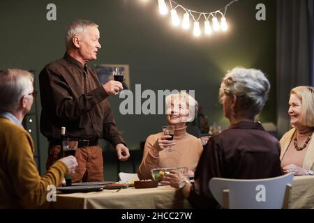 Eine Gruppe älterer Freunde traf sich am Tisch und hatte ein festliches Abendessen. Schöner alter kaukasischer Mann, der mit Glas in der Hand steht und Toast macht Stockfoto