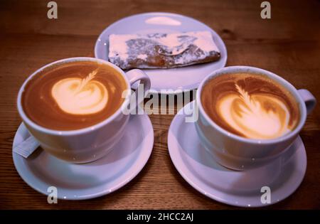 Zwei Cappuccino-Tassen aus weißer Keramik und Kirschkuchen auf einem weißen Keramikteller mit einem Holztisch im Hintergrund Stockfoto