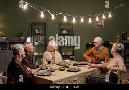 Schöner alter Mann, der Akustikgitarre spielt, während er am Esstisch im dekorierten Wohnzimmer sitzt und mit seinen vier Freunden ein festliches Abendessen hat Stockfoto