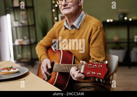 Älterer kaukasischer Mann in legerer Kleidung und Brille sitzt am Esstisch und spielt akustische Gitarre Stockfoto