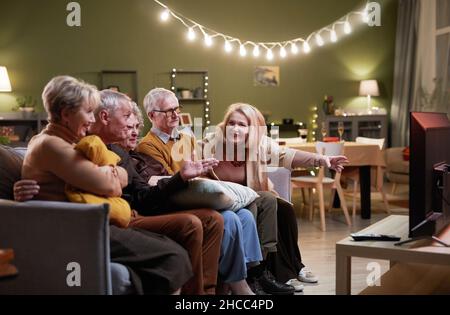 Zwei ältere Männer und drei Frauen sitzen zusammen auf der Couch vor dem Fernseher, schauen sich interessante Filme an und diskutieren darüber Stockfoto