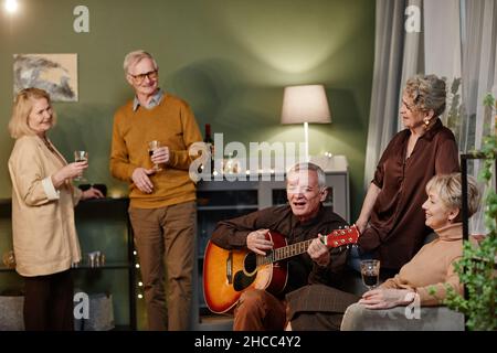 Talentierter Mann im Alter von Kaukasien, der beim Treffen mit Freunden akustische Gitarre spielt und singt. Vier Senioren lauschen ihrer gebratenen Darbietung Stockfoto