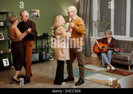 Zwei kaukasische ältere Paare tanzen im mit Lichtern dekorierten Wohnzimmer, während eine ältere Frau auf der Couch sitzt und akustische Gitarre spielt Stockfoto