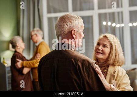 Zwei kaukasische ältere Paare tanzen im Wohnzimmer. Gealterte Frau mit blondem Haar, die beim Tanzen zärtlich auf ihren hübschen Mann schaut Stockfoto