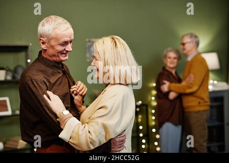 Zwei Paare im Alter, die im beleuchteten Wohnzimmer tanzen. Gealterter, gut aussehender Mann mit grauen Haaren, der beim Tanzen zärtlich auf seine Frau schaut Stockfoto