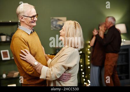 Zwei Paare von Senioren, die im mit Lichtern dekorierten Wohnzimmer tanzen. Der alte kaukasische, gut aussehende Mann trägt eine Brille und blickt zärtlich auf seine Frau Stockfoto