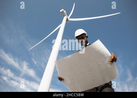 Low-Angle-Ansicht eines fokussierten afroamerikanischen Mannes, der mit Windturbinen auf dem Feld steht und Baupläne untersucht, die in den Händen halten. Konzept für erneuerbare und nachhaltige Energie. Stockfoto