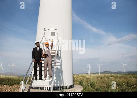 Indischer Ingenieur mit orangefarbenem Helm und grauen Overalls, die in die Ferne zeigen und die Tablette in der Nähe der Windmühle halten. afroamerikanischer Geschäftsmann im Anzug, der bei der Inspektion der Farm in der Nähe stand. Stockfoto