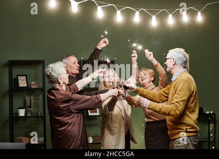 Zwei ältere Männer und drei Frauen stehen zusammen in einem mit Lichtern geschmückten Wohnzimmer, halten Wunderkerzen in den Händen und klirren Gläser während der Festlichkeiten mit mir Stockfoto