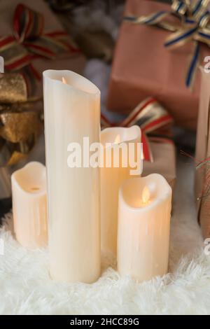 Vier dicke weiße Wachskerzen stehen auf dem weißen, gemütlichen Teppich in der Nähe der Weihnachts-Geschenkschachteln. Geschenke für das neue Jahr, in gewickeltem Papier mit Schleifen. Stockfoto