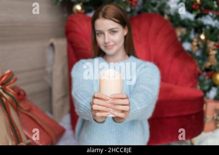 Entzückendes Mädchen sitzt und hält dicke weiße Wachskerze in ihren Händen. Verschwommener Hintergrund, Nahaufnahme, Weihnachtsfeiertage. Stockfoto