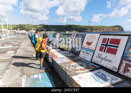 Mutter und Sohn mit einem Rucksack und Blick auf die bemalte Hafenwand mit Graffiti mit dem Berg Pico im Hintergrund, Horta, Insel Faial, Azoren, Portugal Stockfoto