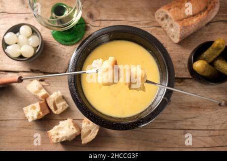 Fondue aus geschmolzenem Käse mit Brot auf langen Gabeln, Gurken und Wein auf einem rustikalen Holztisch, traditionelles Neujahr-Gericht aus der Schweiz, Kopierraum, Stockfoto