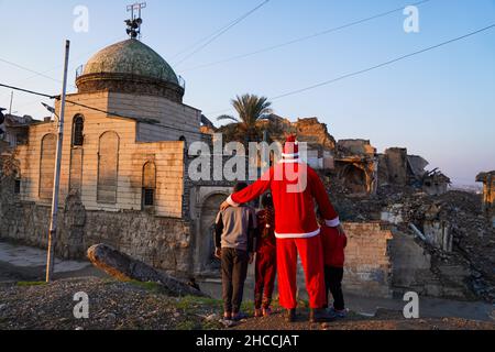 Ein Mann, der als Weihnachtsmann gekleidet ist, steht vor der beschädigten Umayyad-Moschee (Al-Masfe) in der Altstadt von Mosul.eine Initiative irakischer Freiwilliger der Mosul Eye Foundation, um Tausende von Bäumen in der vom Krieg verwüsteten Stadt Mosul zu Pflanzen, Mit dem Ziel, Mosul zu grünen und die Wüstenbildung zu bekämpfen, verteilten sie 100 verschiedene Bäume an die Häuser der Altstadt von Mosul im Rahmen eines Projekts, das darauf abzielte, 5.000 Bäume in der nördlichen Stadt Mosul zu Pflanzen, Das immer noch unter der Zerstörung und Verwüstung leidet, die der Krieg gegen den IS hinterlassen hat. Stockfoto