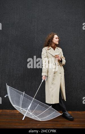 Vertikale Vollaufnahme einer jungen Frau, die einen Trenchcoat mit transparentem Regenschirm und einer Tasse Kaffee trägt, die im Freien gegen eine graue Wand steht und wegschaut Stockfoto