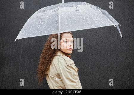 Horizontale mittlere Aufnahme einer schönen jungen Frau mit lockigen Haaren, die einen Trenchcoat trägt und unter einem Regenschirm im Freien gegen eine dunkelgraue Wand steht Stockfoto