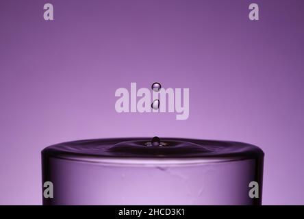 Zwei Tröpfchen fallen auf violettem Hintergrund in ein Glas mit Flüssigkeit Stockfoto