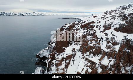 Luftaufnahme von steilen Felsen mit weißem Schnee durch das blaue Meer bedeckt, Schönheit der Winter Natur Konzept. Flug über die wunderschöne felsige Küste in der Wintersaison. Stockfoto