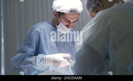 Nahaufnahme von männlichen Chirurgen bei der Arbeit. Seitenansicht konzentrierter Ärzte, die während der Operation auf den Patienten blicken und eine schützende medizinische Uniform tragen Stockfoto