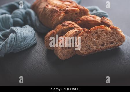 Hausgemachte, frisch gebackene, köstliche Baguette aus Getreidemehl auf schwarzem Hintergrund mit grauer Gaze-Tischdecke Stockfoto