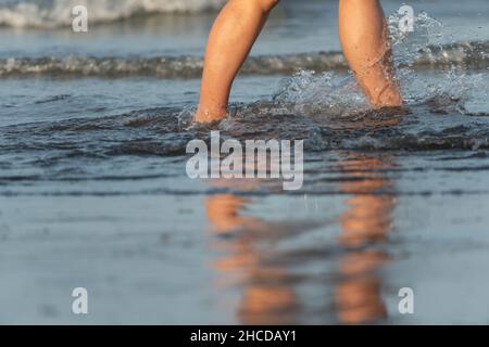 Schöne Beine einer jungen Frau, die an einem Strand mit Wasser, das gegen ihre Füße spritzt, läuft Stockfoto