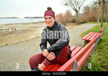 Lindau Am Bodenseee, Deutschland. 16th Dez 2021. Die Athletin Valeria kleiner sitzt auf einer Parkbank am Ufer des Bodensees. Im Hintergrund ist die Stadt Lindau zu sehen. Kleiner spielte in der DFB-Auswahl, gewann Titel, spielte in der Frauen-Fußball-Bundesliga. Verletzungen verursachten eine Veränderung. Jetzt tritt sie als Triathletin an. (Zu 'Triathlon nach dem Fußball: Faszination und Härten einer professionellen Karriere von 2nd') Quelle: Felix Kästle/dpa/Alamy Live News Stockfoto