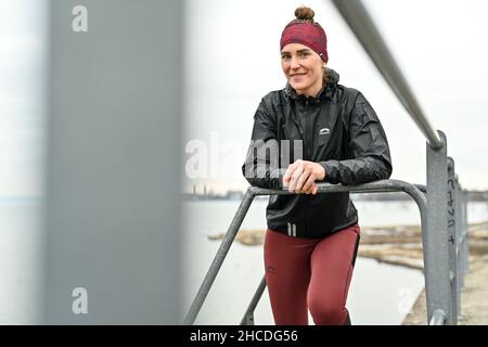 Lindau Am Bodenseee, Deutschland. 16th Dez 2021. Die Athletin Valeria kleiner sitzt auf einer Parkbank am Ufer des Bodensees. Im Hintergrund ist die Stadt Lindau zu sehen. Kleiner spielte in der DFB-Auswahl, gewann Titel, spielte in der Frauen-Fußball-Bundesliga. Verletzungen verursachten eine Veränderung. Jetzt tritt sie als Triathletin an. (Zu 'Triathlon nach dem Fußball: Faszination und Härten einer professionellen Karriere von 2nd') Quelle: Felix Kästle/dpa/Alamy Live News Stockfoto