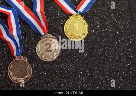 Gold-, Silber- und Bronzemedaillen mit den Nummern eins, zwei, drei und Bändern auf schwarzem Glitzer-Hintergrund. Sporttrophäe. Konzept für Sieg oder Erfolg, Award-Konzept Stockfoto