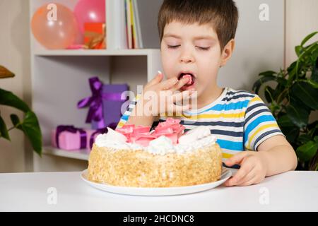 Ein glücklicher Junge isst einen Geburtstagskuchen, isst mit den Händen und leckt sich die Finger. Stockfoto