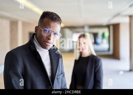 Afroamerikanischer Mann in stilvollem Mantel und Brille, der die Kamera anschaut, während er in einer modernen Passage in der Nähe einer Freundin steht Stockfoto