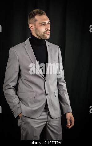 Porträt eines jungen Geschäftsmannes in grauem Anzug und schwarzem Rollkragen, ernst aussehend mit einer Hand in den Taschen Stockfoto