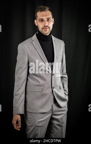 Porträt eines jungen Geschäftsmannes in grauem Anzug und schwarzem Rollkragen, ernst aussehend mit einer Hand in den Taschen Stockfoto
