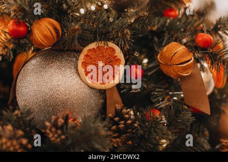 Getrocknete Orangen, Beeren, Zapfen, die am Weihnachtsbaum hängen. Kreative festliche Details und goldener Weihnachtsball. Winterzauber im Neujahrsstil Stockfoto