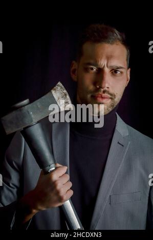 Der junge Geschäftsmann mit grauem Anzug, der eine Axt in der Hand hält, hat ein entschlossenes und bedrohliches Aussehen - ein aggressives Trading-Methodenkonzept Stockfoto