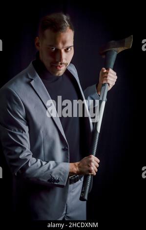 Der junge Geschäftsmann mit grauem Anzug, der eine Axt in der Hand hält, hat ein entschlossenes und bedrohliches Aussehen - ein aggressives Trading-Methodenkonzept Stockfoto