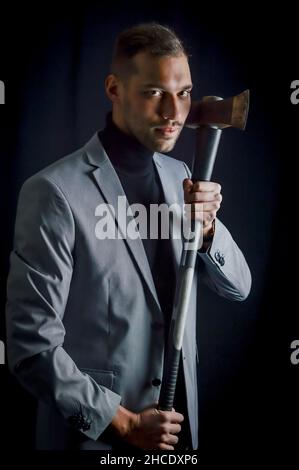 Der junge Geschäftsmann mit grauem Anzug, der eine Axt in der Hand hält, hat ein entschlossenes und bedrohliches Aussehen - ein aggressives Trading-Methodenkonzept Stockfoto