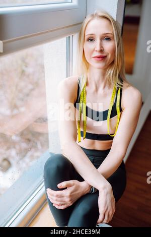 Junge schöne sportliche blonde Frau in schwarz-rosa Oberteil und schwarzen Leggings auf Fensterbank mit Maßband am Hals sitzen. Frau Sportswear concep Stockfoto