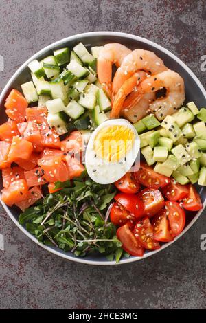 Köstlicher gesunder Salat aus Lachs, Garnelen, Eiern, Avocado, Gurken und Tomaten aus nächster Nähe auf einem Teller auf dem Tisch. Vertikale Draufsicht von oben Stockfoto