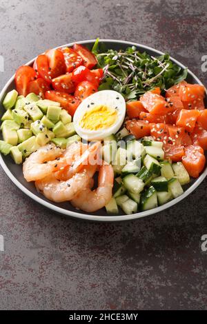 Köstlicher gesunder Salat aus Lachs, Garnelen, Eiern, Avocado, Gurken und Tomaten in Nahaufnahme auf einem Teller auf dem Tisch. Vertikal Stockfoto