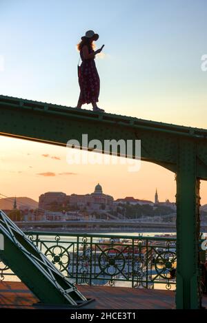 Junge Frau, die auf dem Brückenbalken läuft und ihr Telefon gegen die Stadt anstarrt Stockfoto